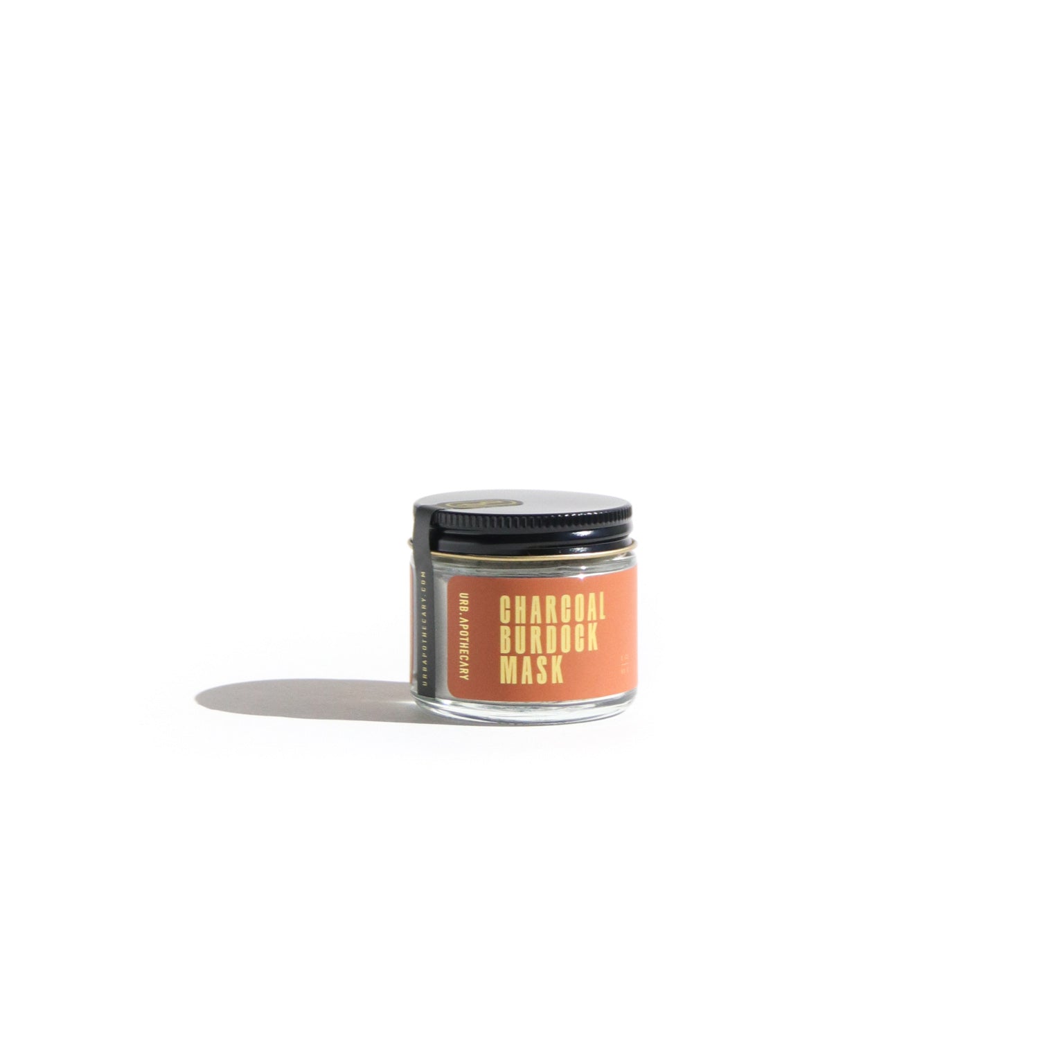 Minimalist product photo of Urb Apothecary Charcoal Burdock Mask in clear glass jar with black metal lid and bold orange label, natural clay face mask for detoxifying and calming irritated skin