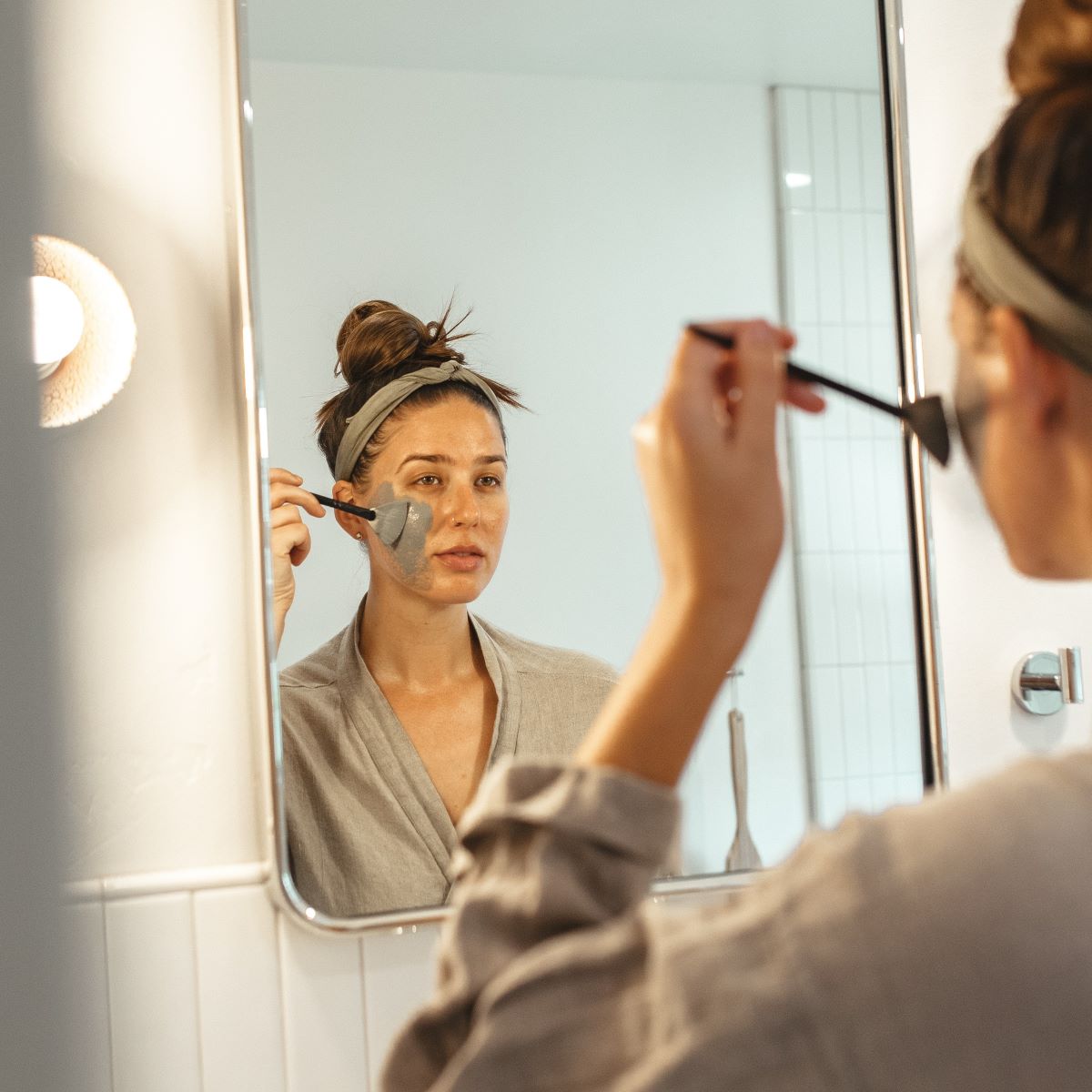 Woman applying Urb Apothecary Charcoal Burdock Mask in front of bathroom mirror using a mask brush , natural skincare ritual for detoxifying and soothing breakout-prone skin