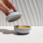Hand lifting lid off speckled ceramic bonbonnière revealing Urb Apothecary’s golden Pot of Gold Regenerative Face Balm, designed to protect, brighten, and deeply nourish sun-exposed skin